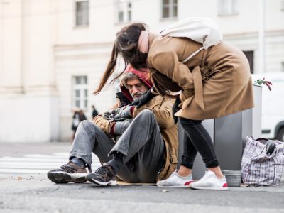 A young woman giving money to homeless beggar man sitting outdoors in city.